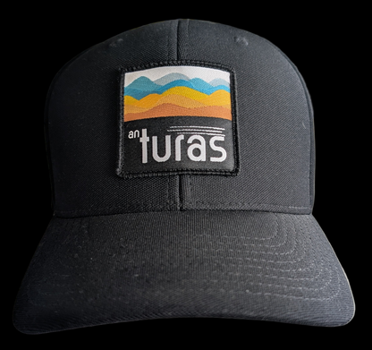 The Sòlas Cap. Black outdoor performance cap with a colorful landscape and An Turas™ logo on a woven patch. Front view.