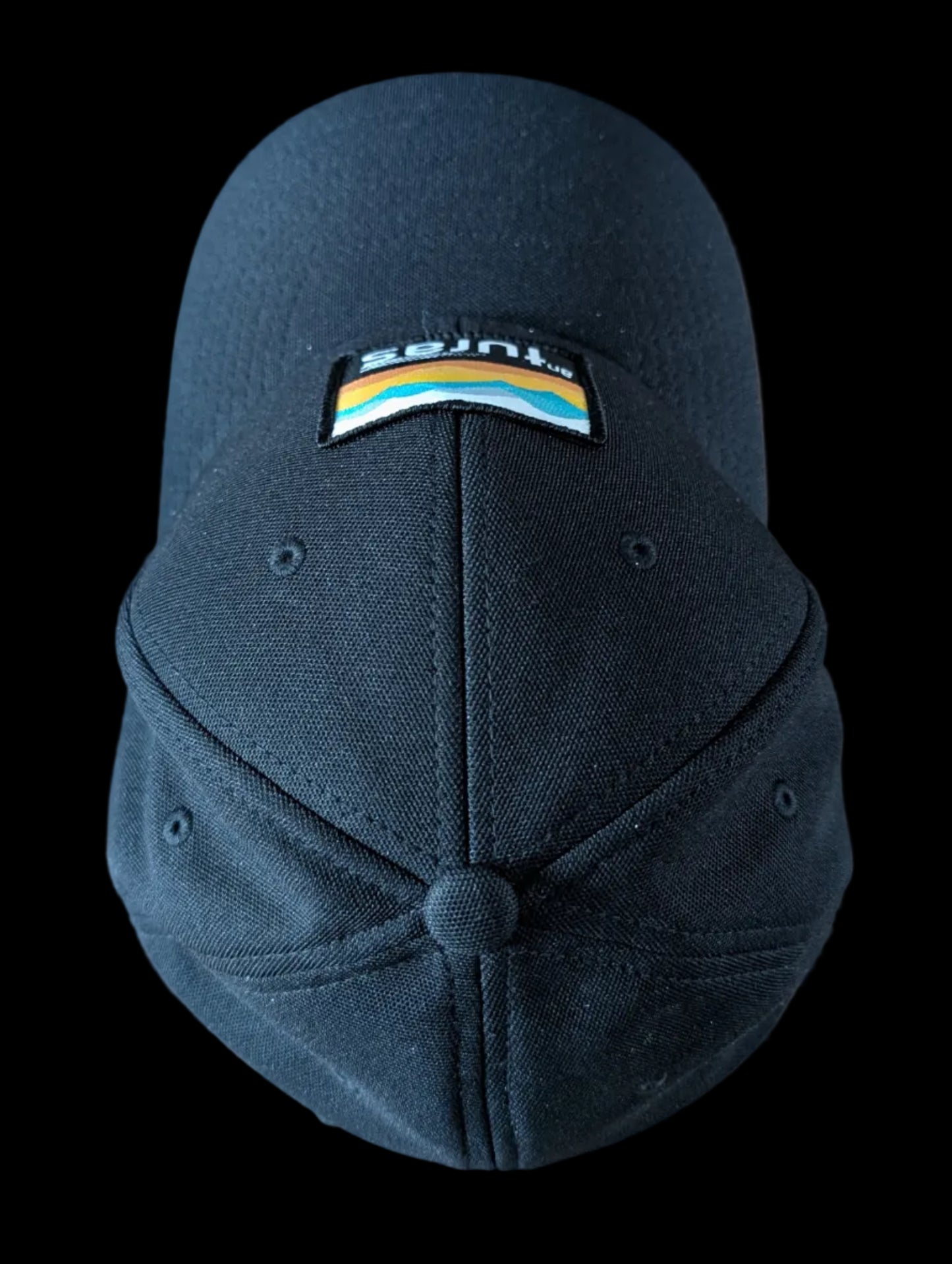 The Sòlas Cap. Black outdoor performance cap with a colorful landscape and An Turas™ logo on a woven patch. Aerial view.