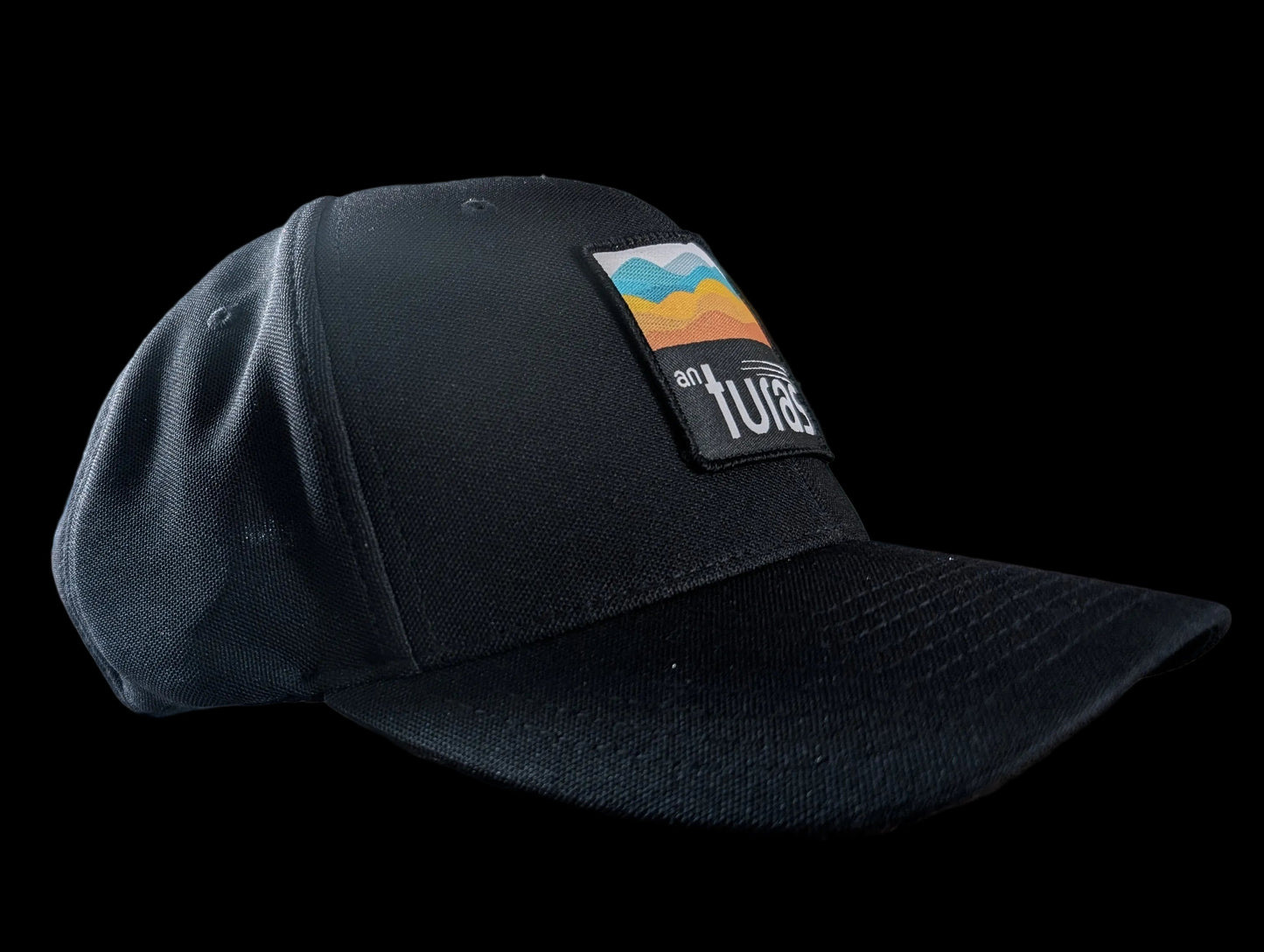 The Sòlas Cap. Black outdoor performance cap with a colorful landscape and An Turas™ logo on a woven patch.